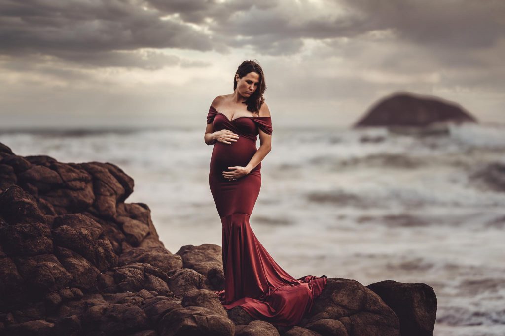 pregnant lady in red dress on beach standing on rocks - Maternity photography Auckland - Amber Farooq