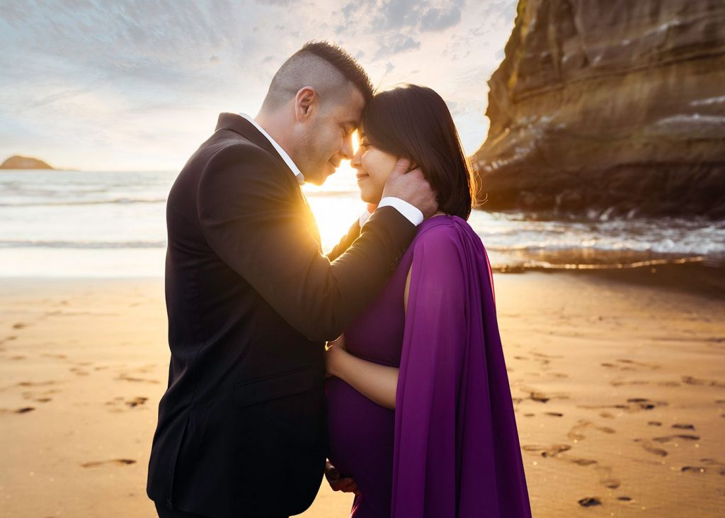 pregnant wife and husband hugging Auckland Maternity Photography Session at Bethells Beach during sunset