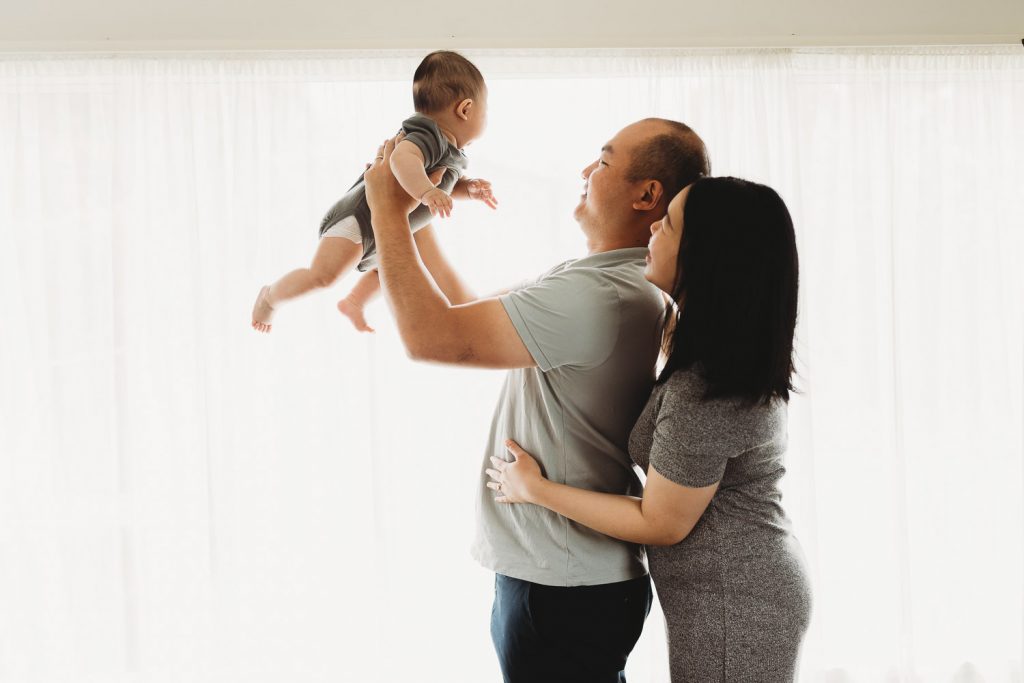 mother father holding newborn and standing beside window - Auckland Lifestyle Newborn Photographer Amber Farooq