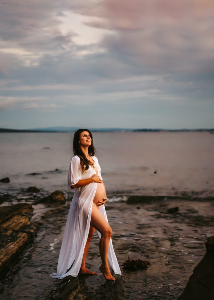Pregnant woman in a flowing white dress, holding her baby bump in a field during sunset