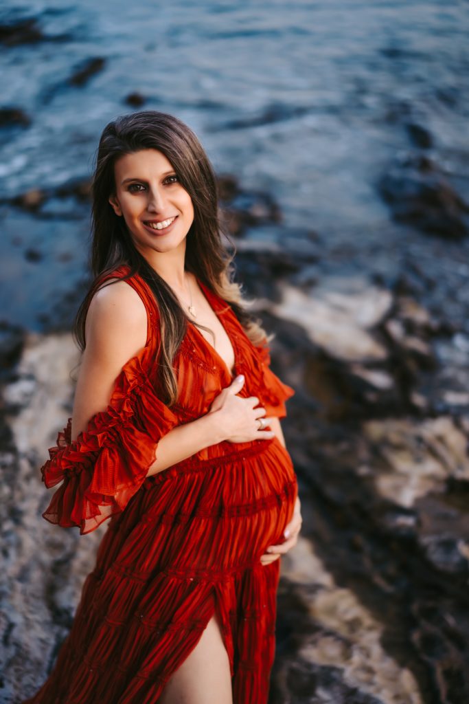 Radiant pregnant woman in a beautiful red dress standing on the beach, enjoying the picturesque view and the company of her baby bump