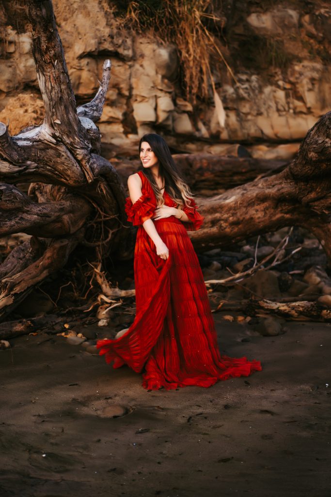 Pregnant woman in a stunning red dress standing on the beach with her baby bump on display, feeling the cool breeze on her face