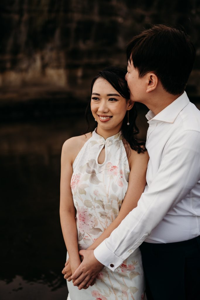 Lishan playfully dips Ernest amidst the black sand dunes of Muriwai Beach, celebrating their honeymoon with a touch of fun.