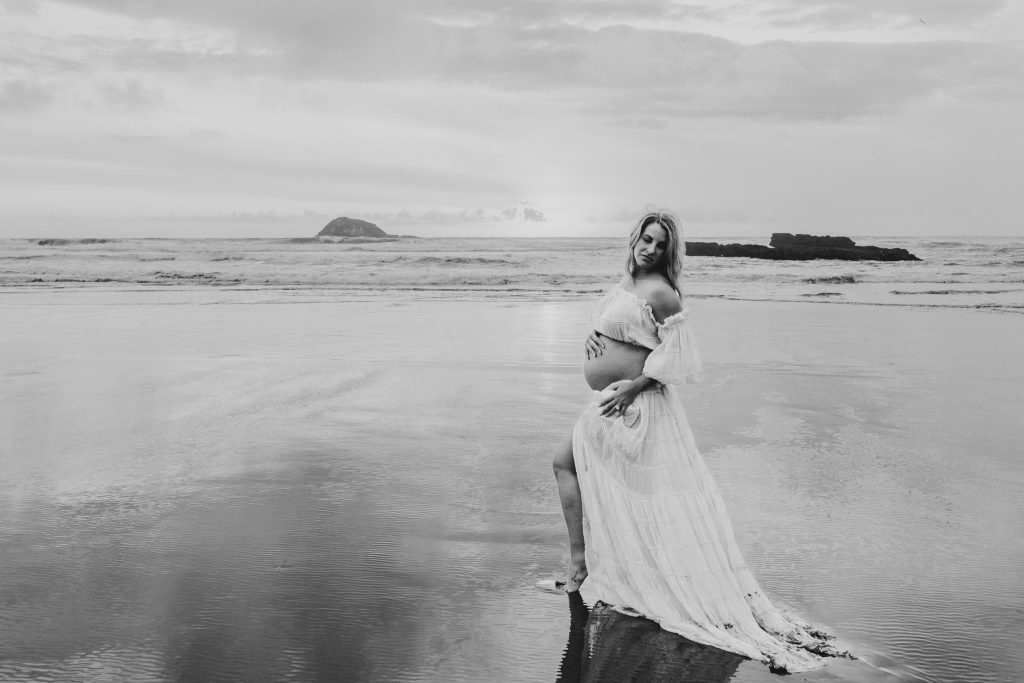 Auckland maternity photo: Mom-to-be cradles baby bump on scenic Auckland beach.