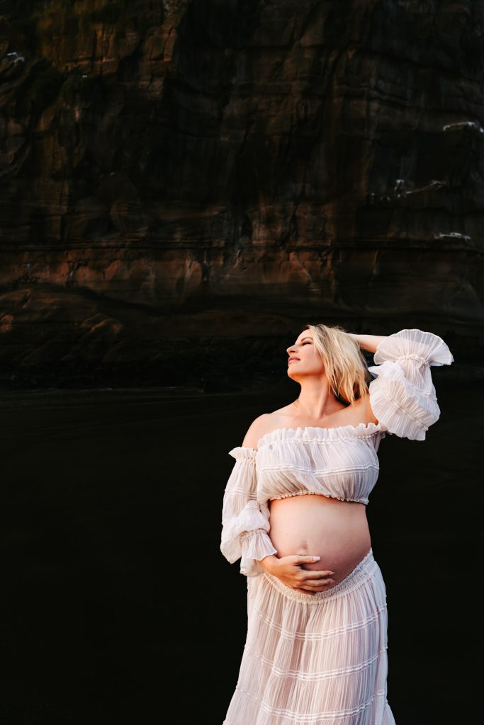 Auckland maternity photography: Sunlight bathes a pregnant woman in a flowing dress, captured by an Auckland maternity photographer.